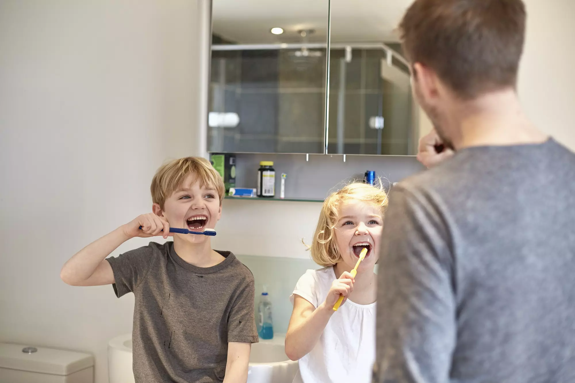 Two children and an adult man cleaning their teeth with toothbrushes in a bathroom.